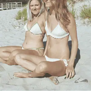 Two women wearing handmade crochet bikinis sitting on a sandy beach.