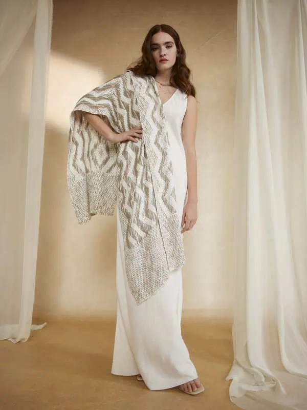 Model wearing a flowing white dress and patterned shawl made from organic cotton in a minimalist setting.