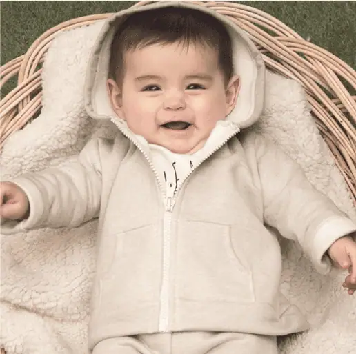 Smiling baby in a cream-colored organic cotton hoodie lying in a wicker basket with fleece lining.