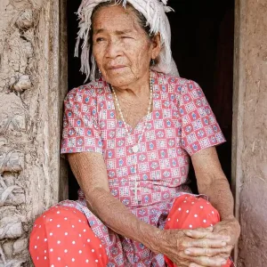 Elderly woman sitting in a doorway of a rustic adobe home, wearing traditional attire.