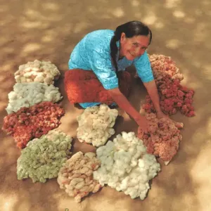 Smiling woman sitting on the ground with piles of naturally colored cotton in various shades surrounding her.