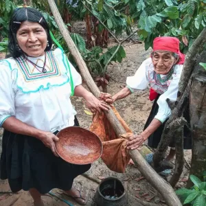 Two Andean artisan women welcoming guests during textile sourcing experiences Peru while filtering liquid dye through a cloth in a traditional jungle setup.